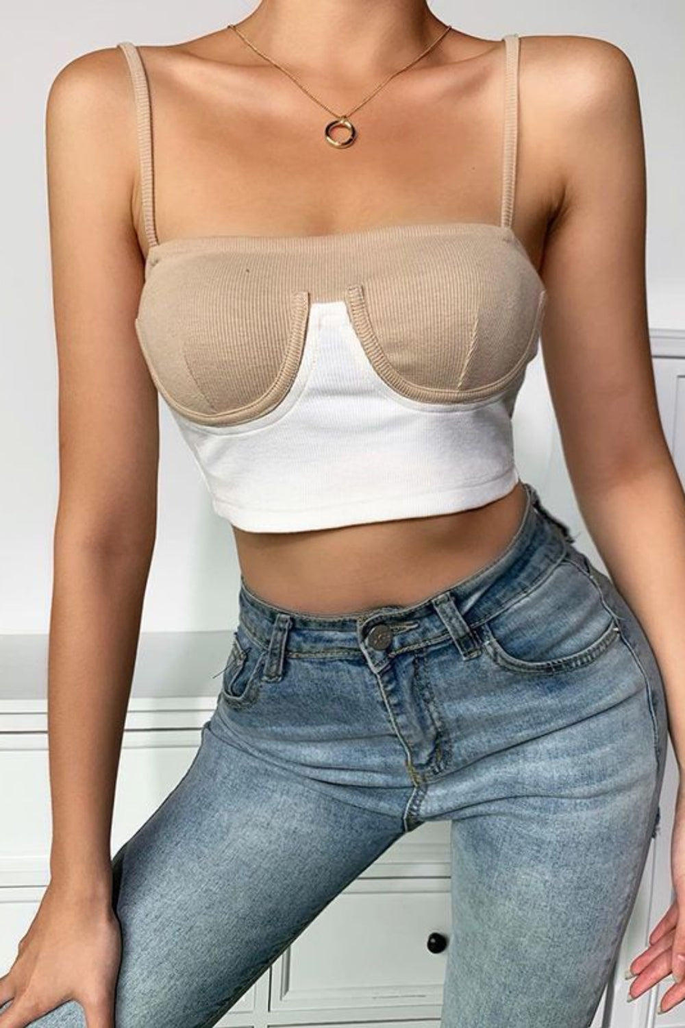 Person wearing a beige bralette over a white crop top and blue jeans indoors.