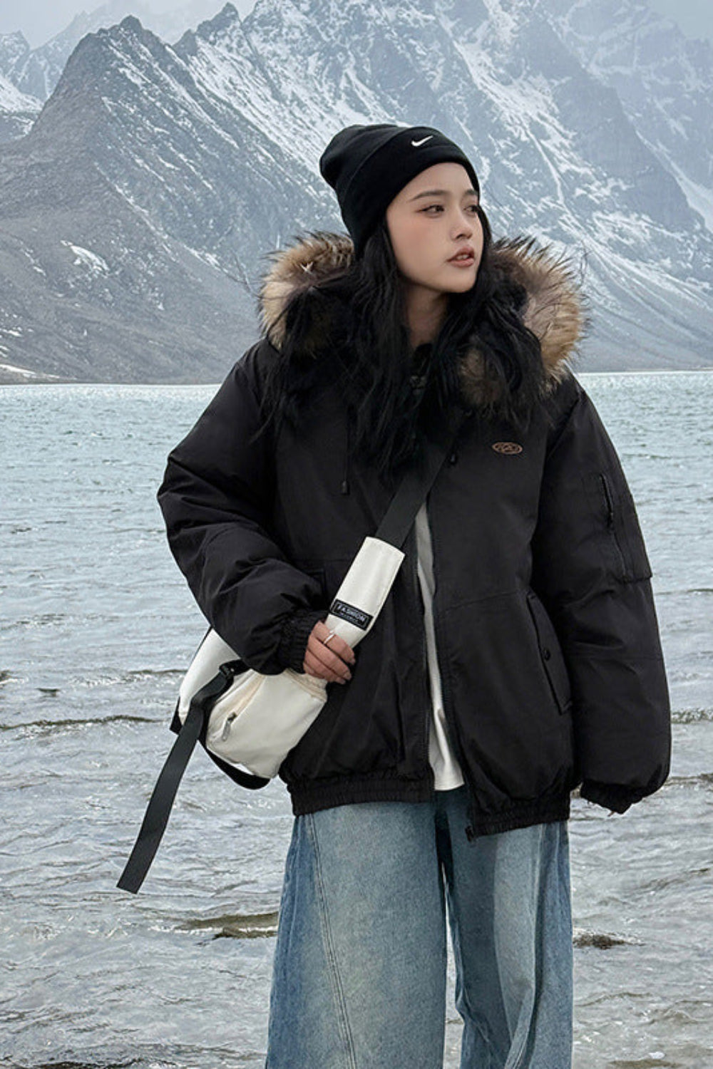 Person wearing a black coat with fur hood standing by a lake with mountains in the background