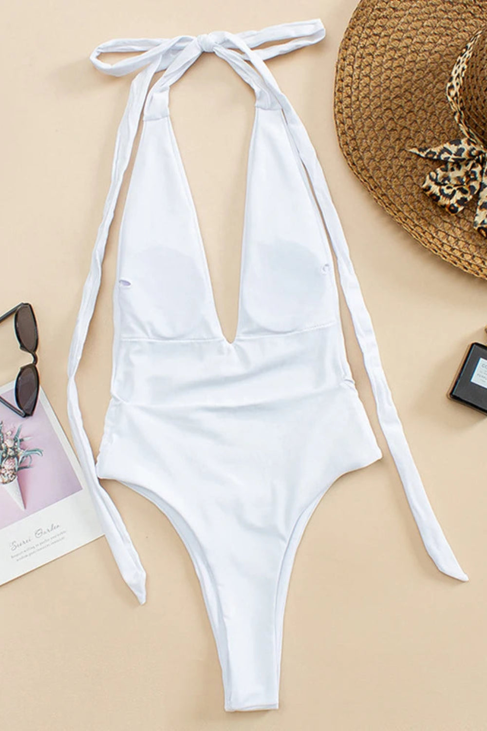 White swimsuit with shoulder ties on a beige background with beach accessories.
