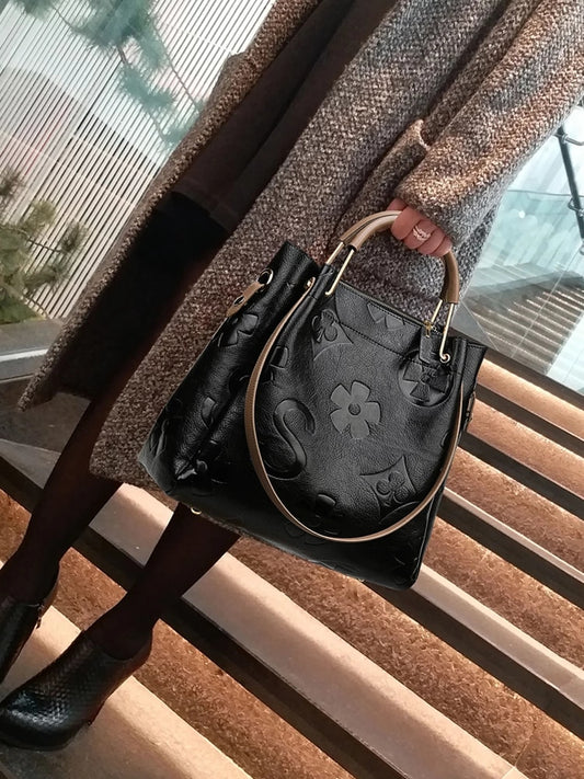 A woman's black embossed handbag with a structured design being held by a person, featuring a hasp closure.