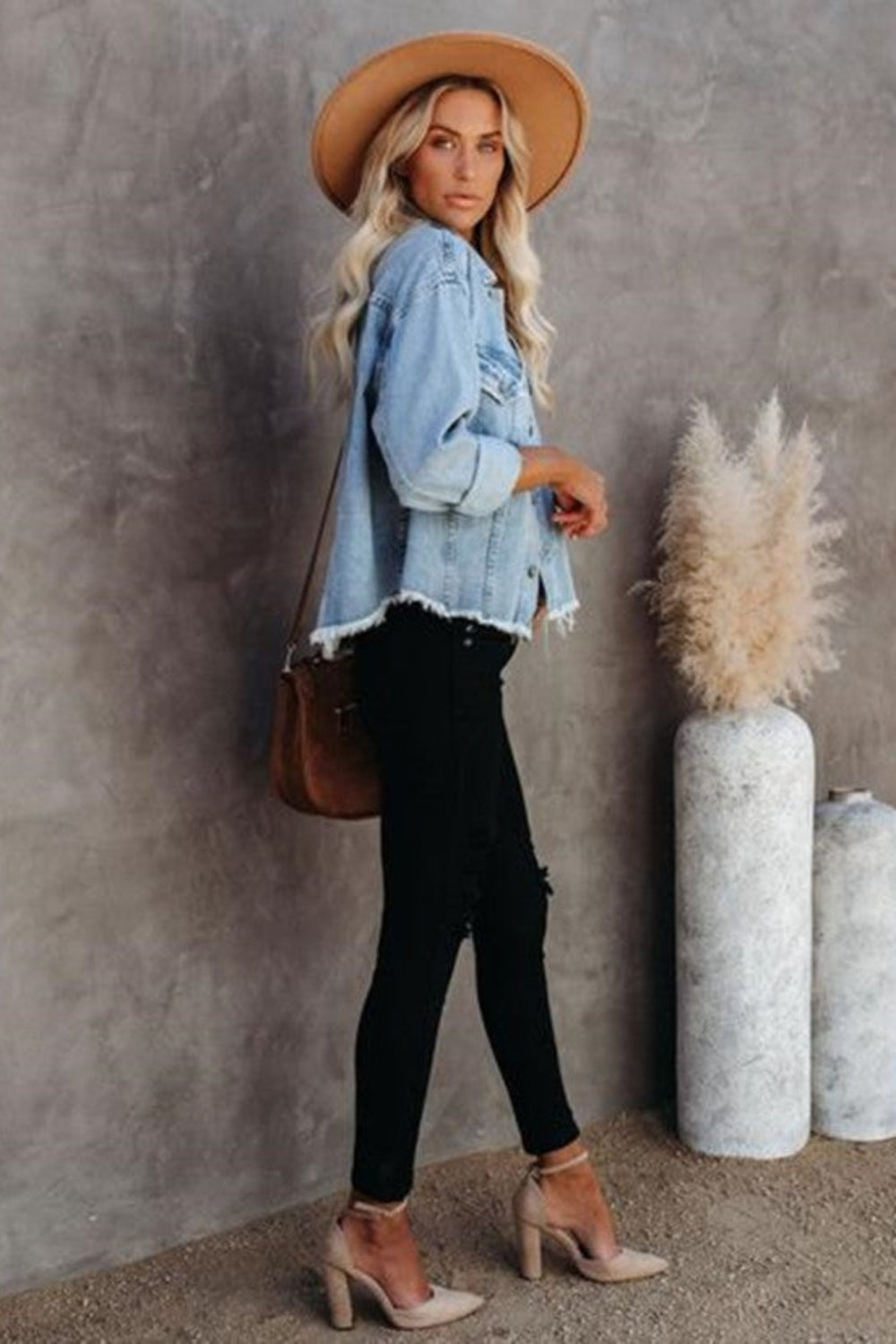 Woman in denim jacket and black pants standing against a gray wall with decorative vases.
