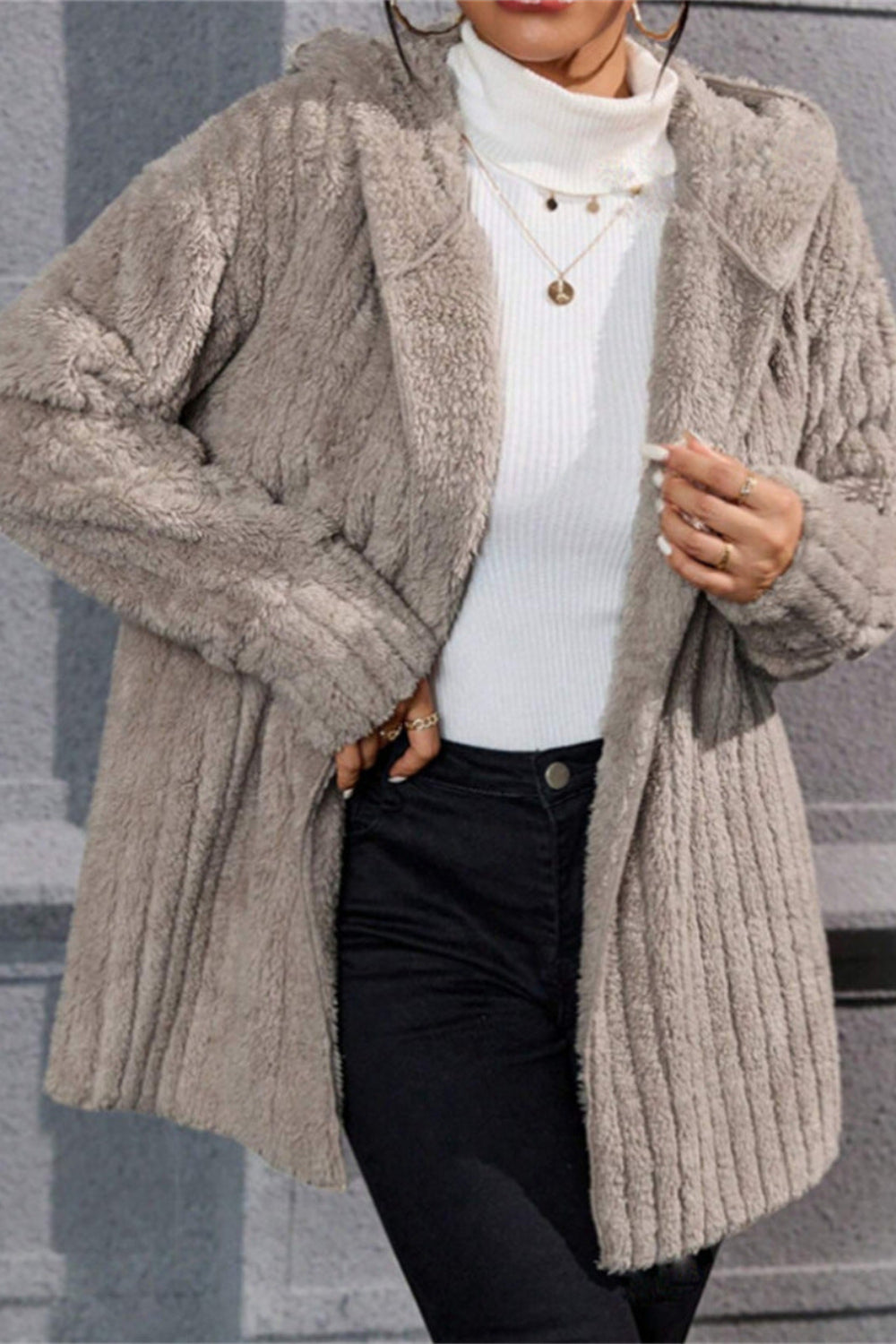 Person wearing a beige knitted cardigan over a white turtleneck and black pants, standing against a gray wall.