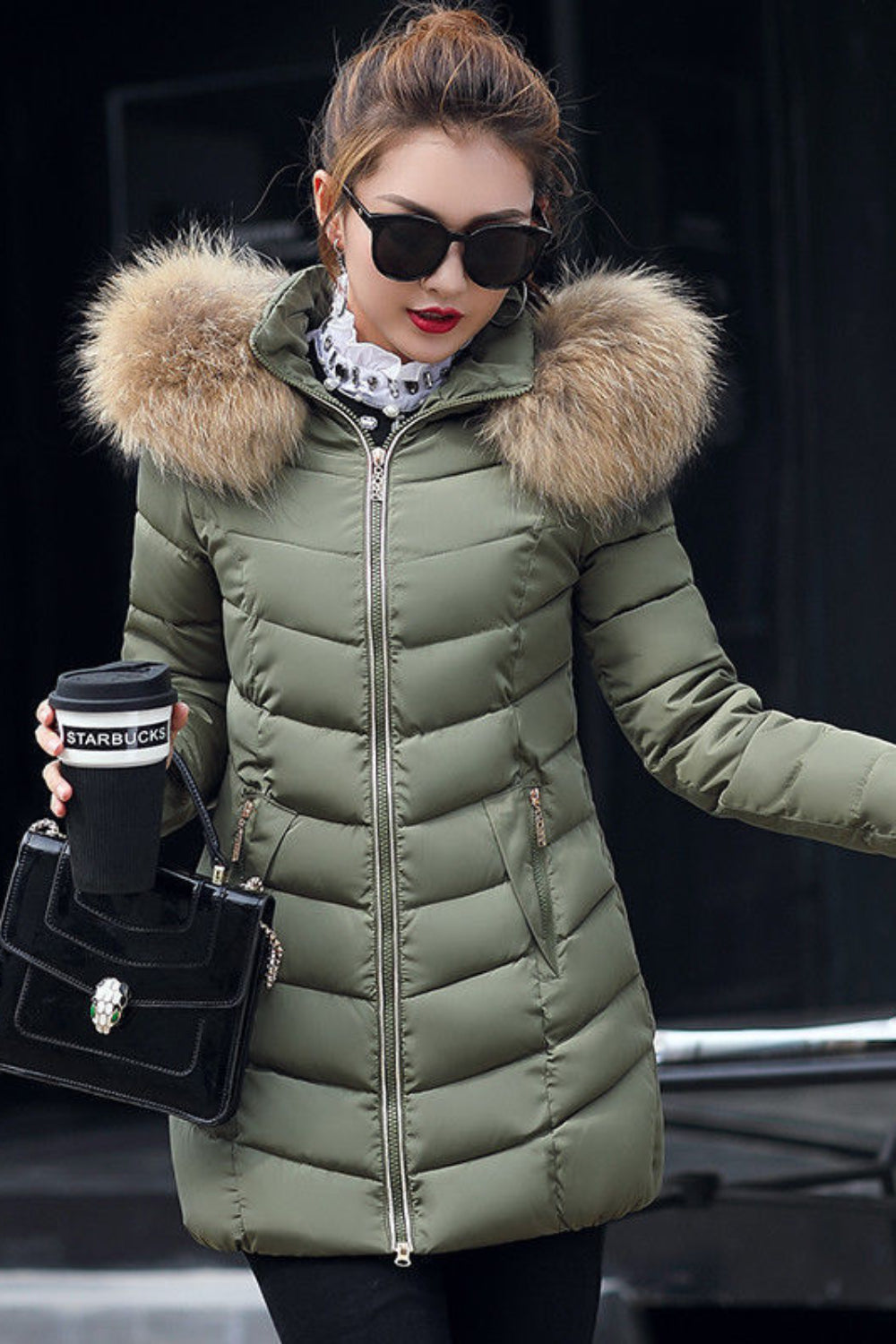 Woman wearing a green puffer jacket with fur hood, holding a coffee cup and black handbag.