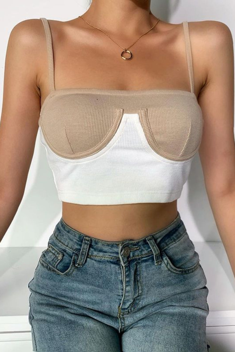 Person wearing a beige bralette over a white crop top and blue jeans on a white background