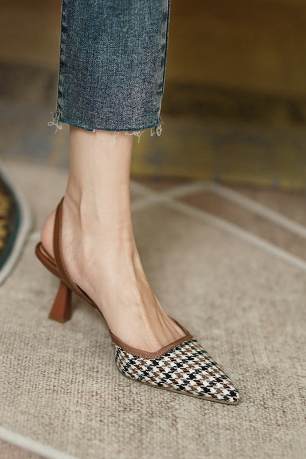 Checkered high-heeled shoes worn with rolled-up jeans on a textured floor.
