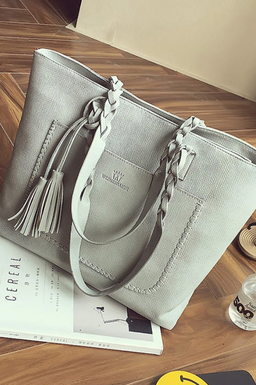 Gray handbag with braided handle on a wooden floor with a magazine and bottle.