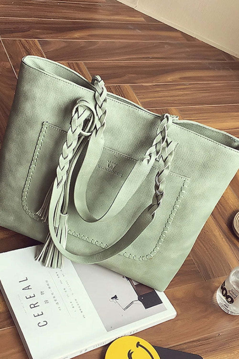 Green handbag with braided handles on a wooden floor with a magazine and bottle.