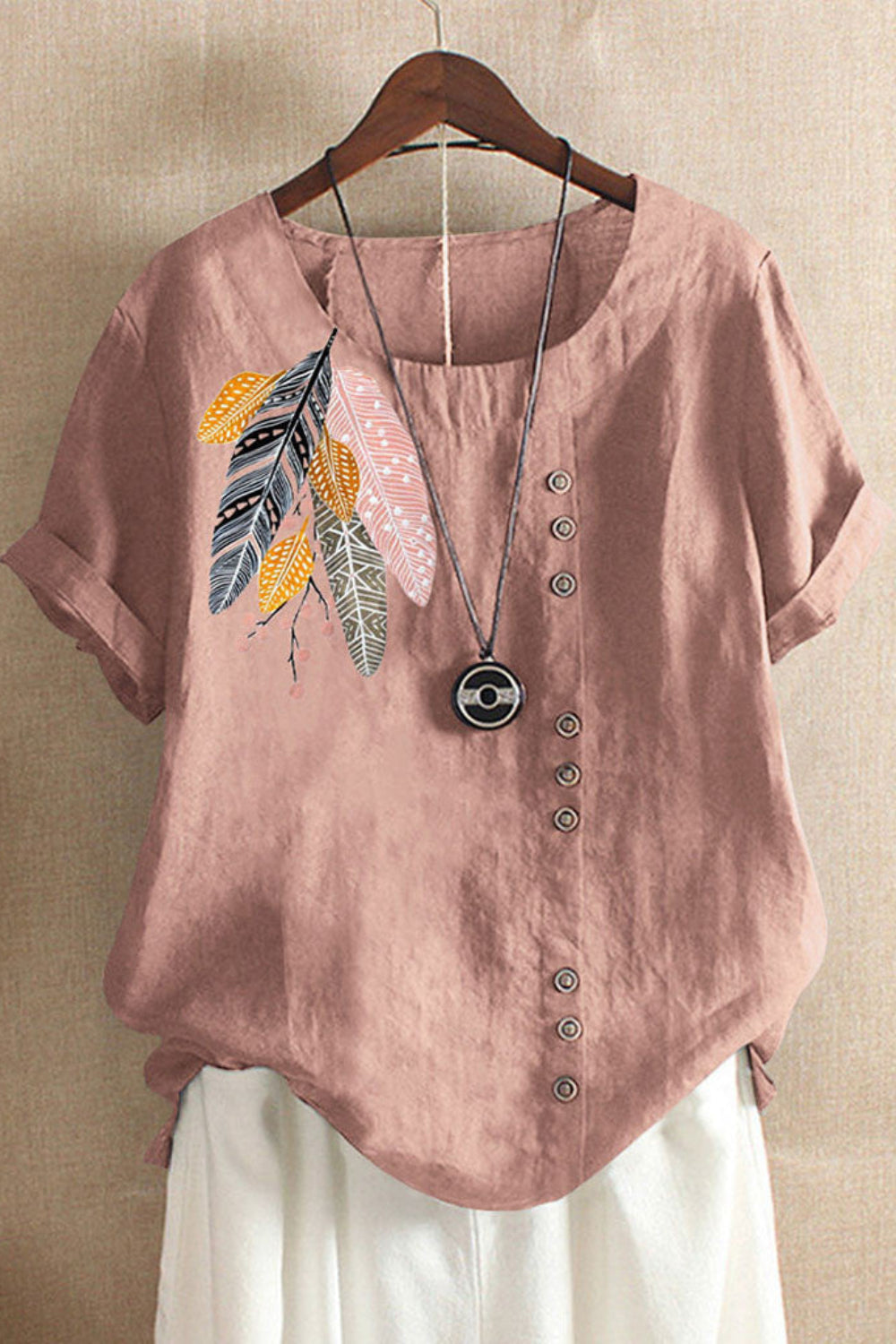 Pink blouse with feather design and buttons on a wooden hanger against a beige wall.