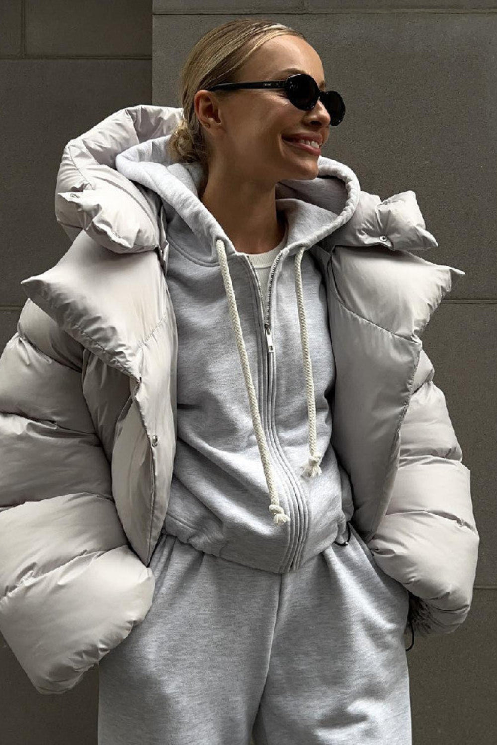 Person wearing a gray puffer jacket and matching outfit against a gray wall.