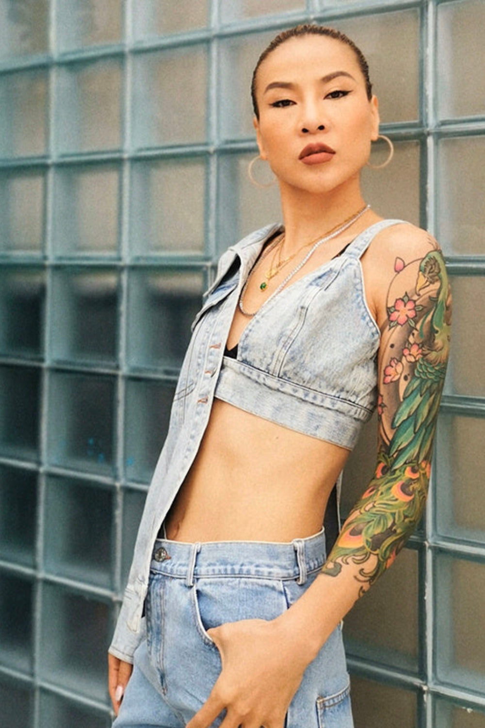 Woman with tattoos wearing a denim outfit against a glass block wall.