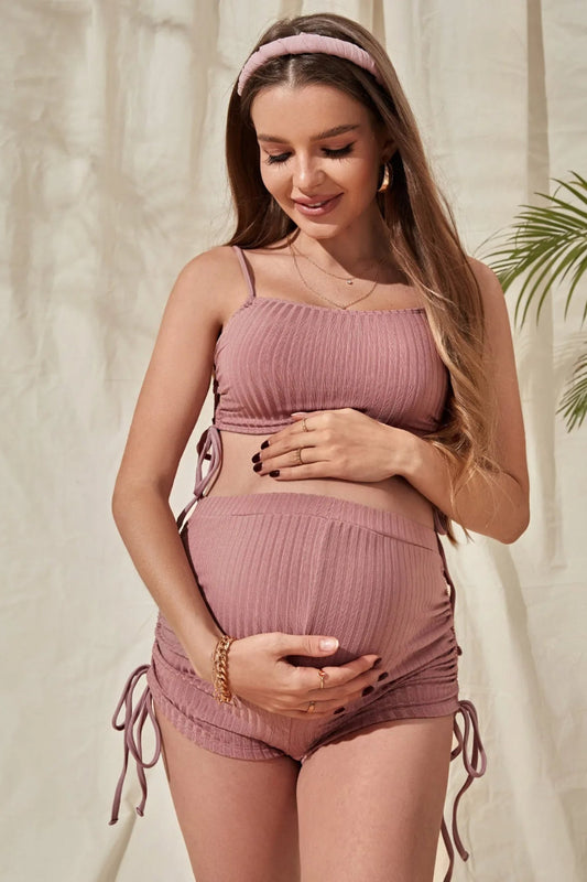 A pregnant woman wearing a pink maternity one-piece swimsuit with side drawstrings. She is also wearing a matching pink headband and has her hand on her belly.
