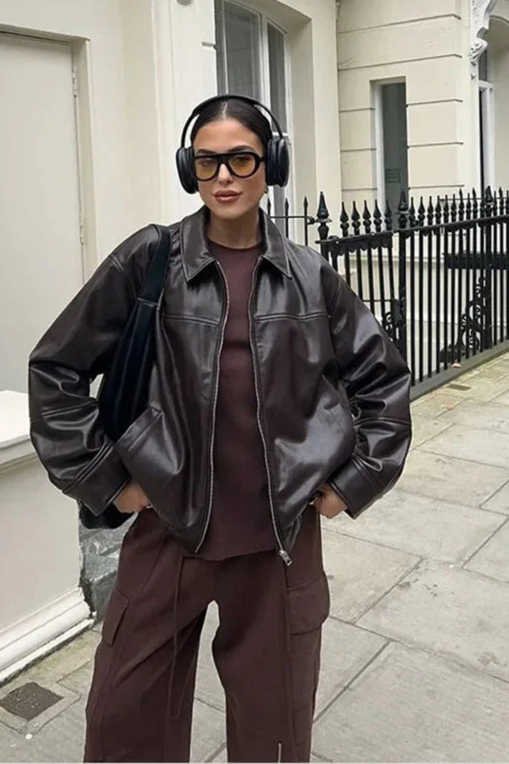 Person wearing a black leather jacket and brown outfit with headphones, standing on a street.
