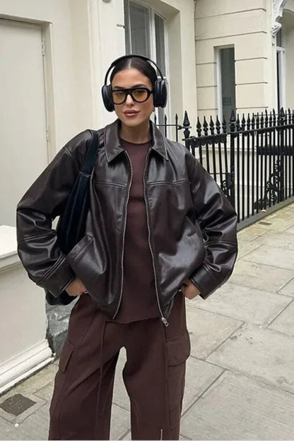 Person wearing a black leather jacket and brown outfit with headphones, standing on a street.