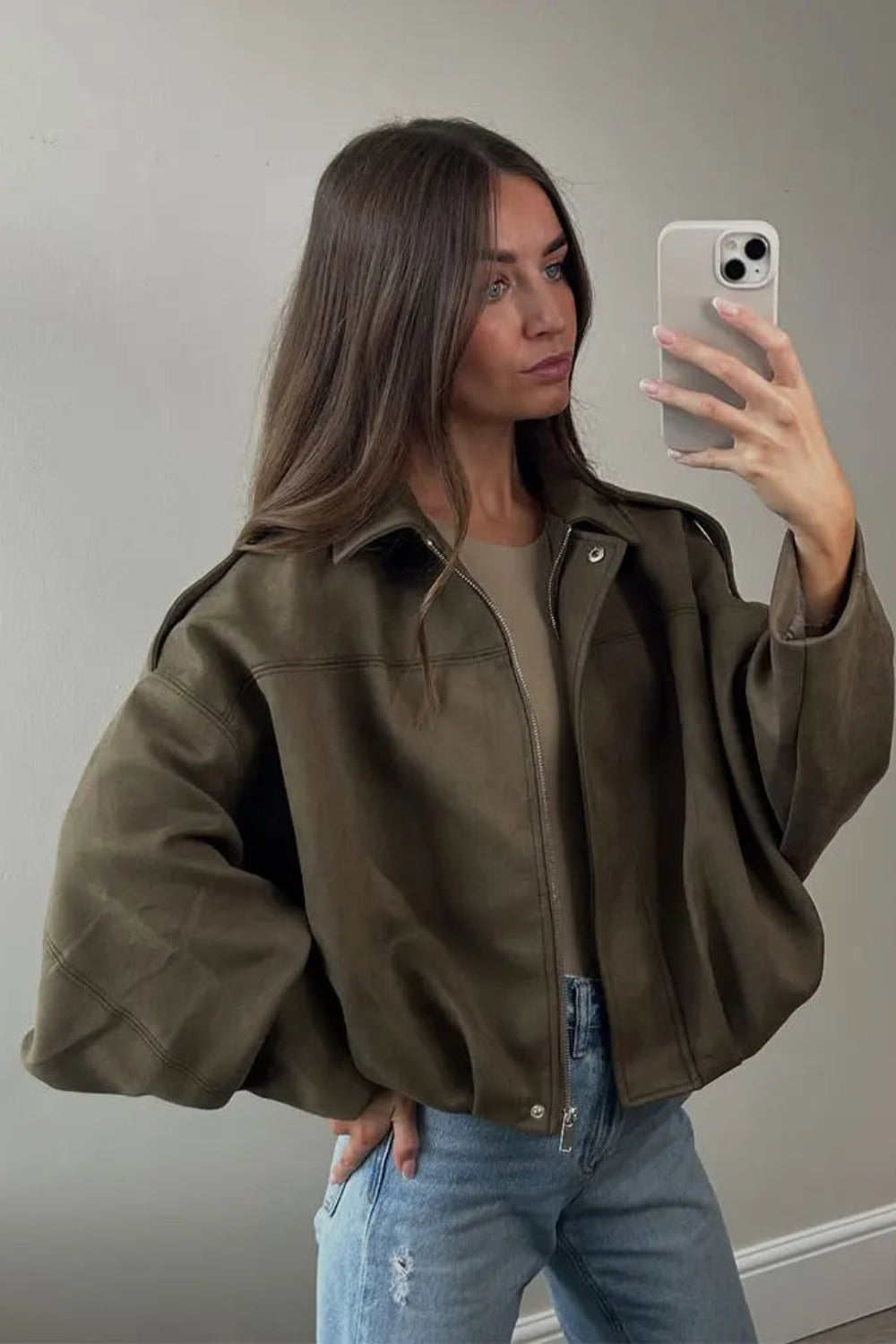 Woman taking a mirror selfie wearing an olive green jacket and light blue jeans.