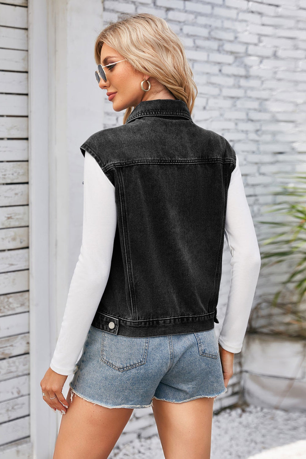 Woman wearing a black denim vest over a white long-sleeve shirt and denim shorts, standing against a light stone wall.