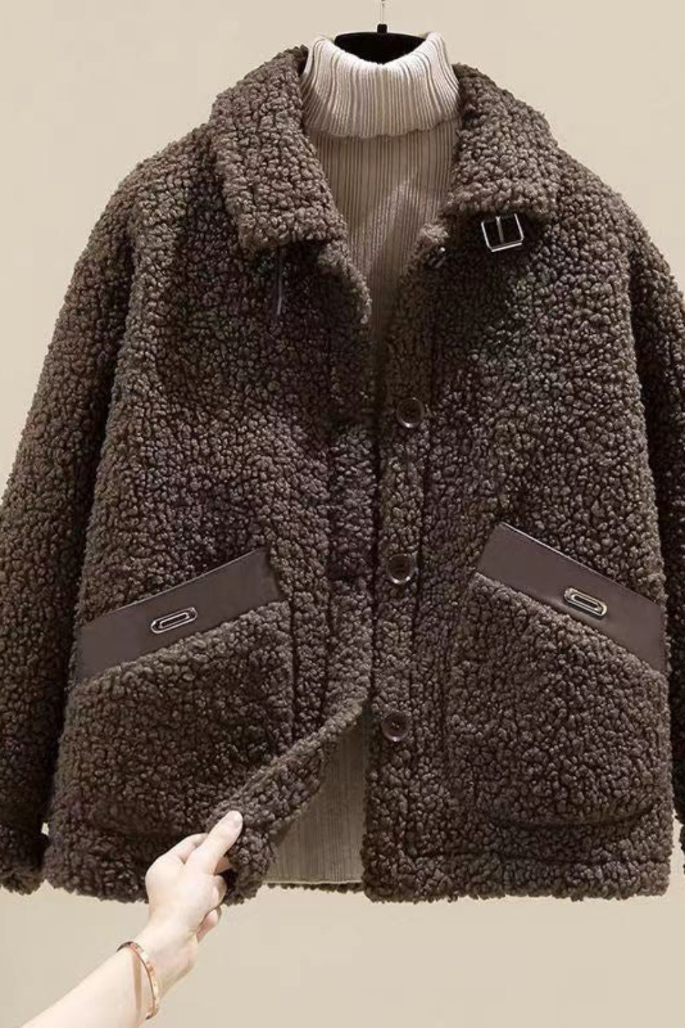 Brown sherpa jacket with a hand holding it against a beige background