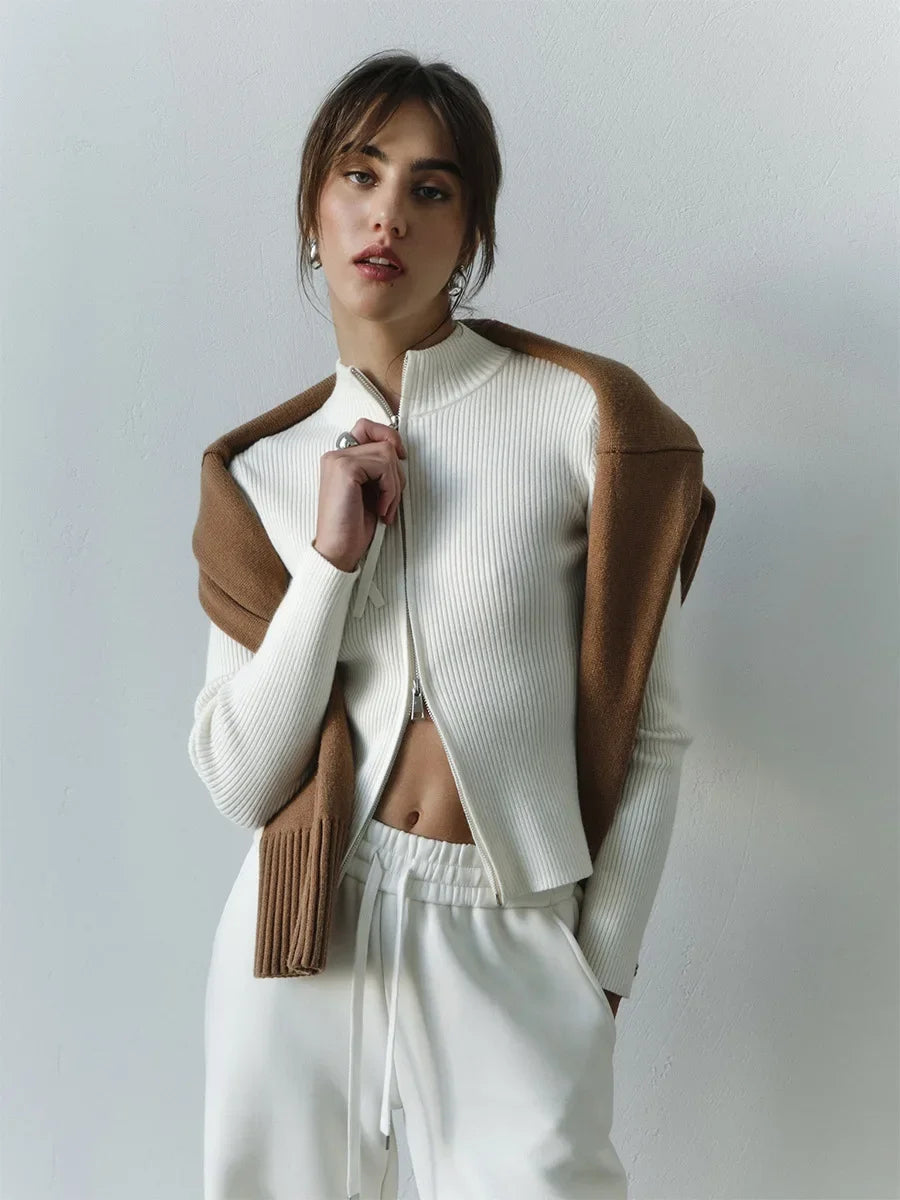 Woman wearing a white ribbed sweater with a brown leather jacket over it, standing against a plain background.