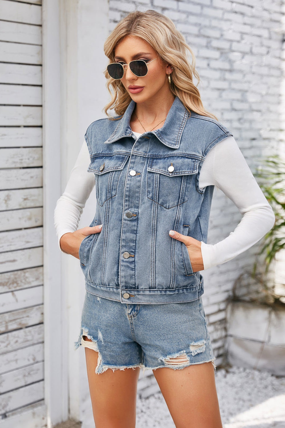 Woman wearing a denim vest and shorts with sunglasses, standing against a light-colored wall.