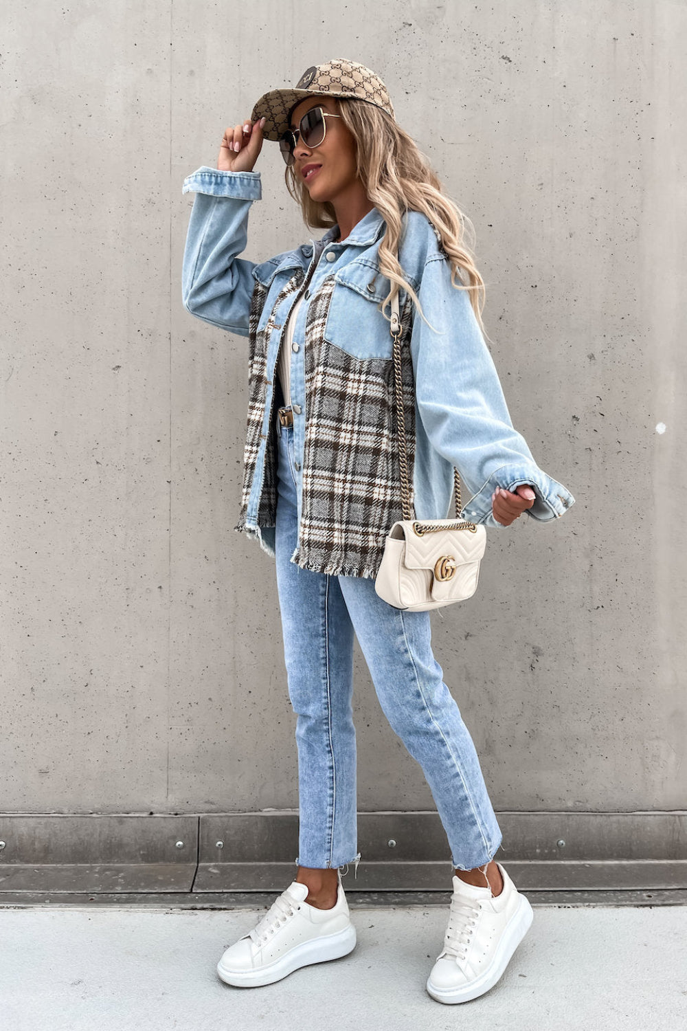 Woman wearing a denim jacket, plaid shirt, and jeans against a gray wall.