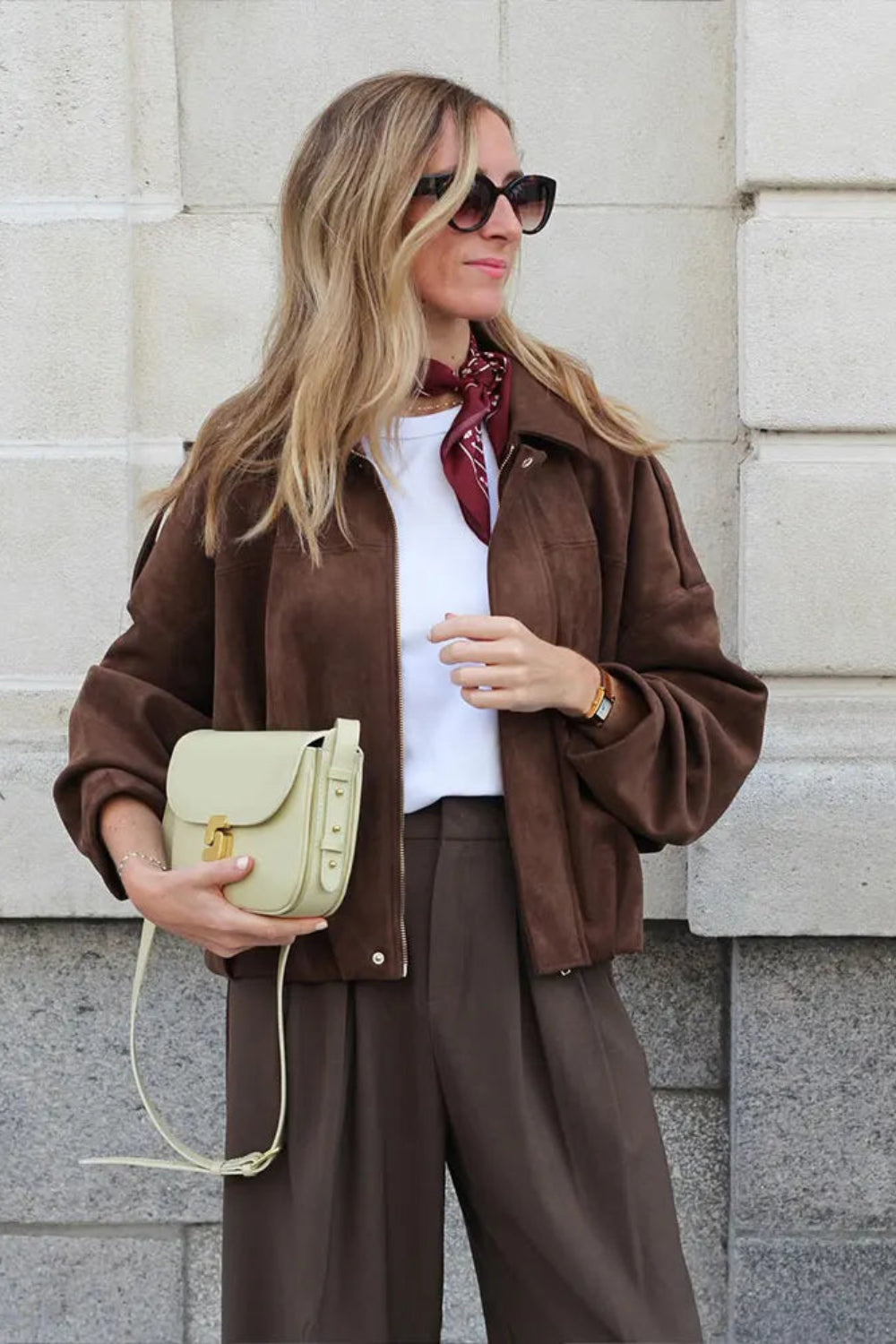 Woman in brown jacket and pants holding a beige handbag against a light stone wall.