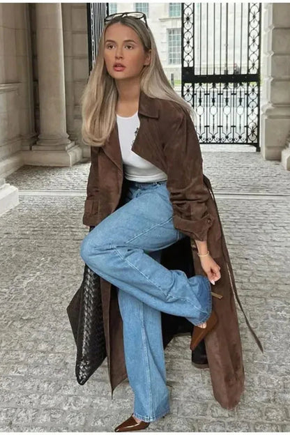 Woman in a brown coat and blue jeans posing in an elegant outdoor setting.