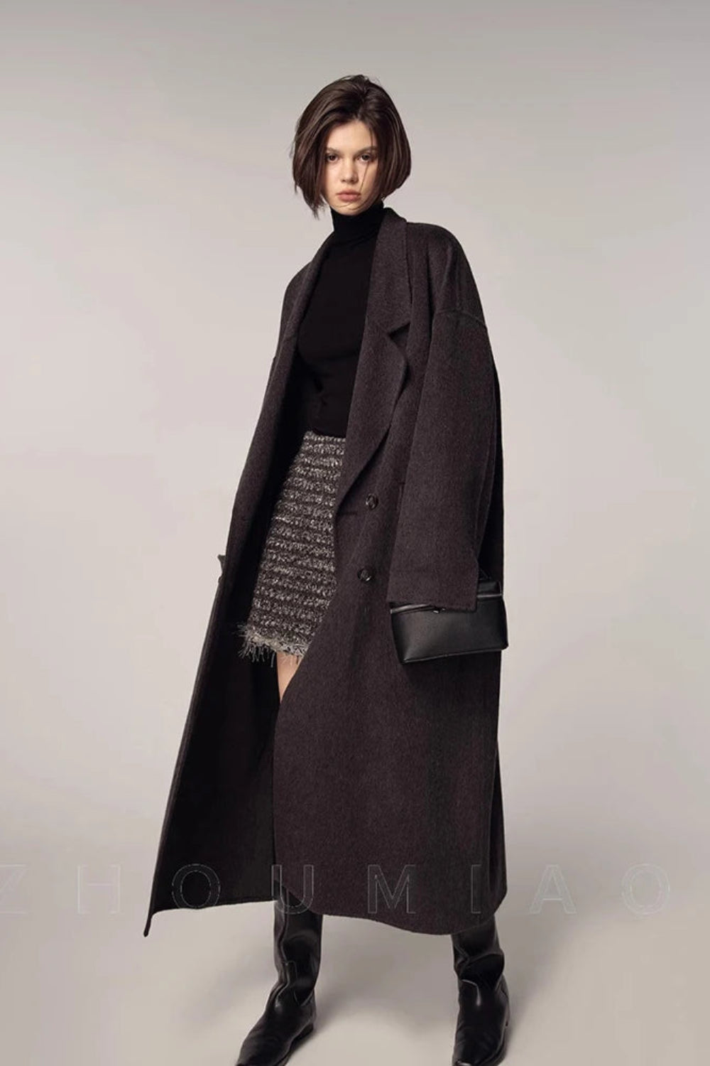 Woman wearing a long dark coat over a black turtleneck and patterned skirt on a plain background