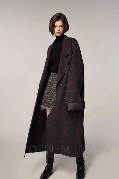 Woman wearing a long dark coat over a black turtleneck and patterned skirt on a plain background