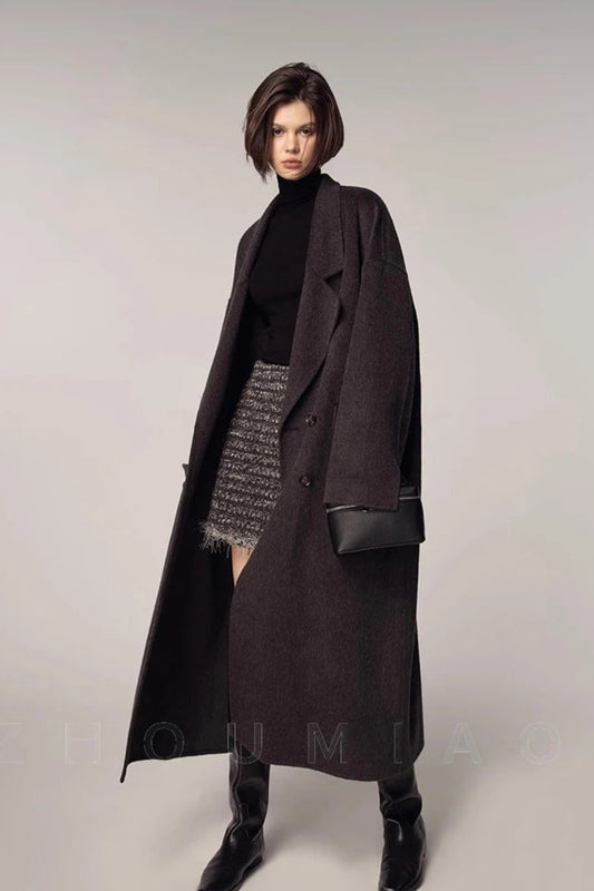 Woman wearing a long dark coat over a black turtleneck and patterned skirt on a plain background