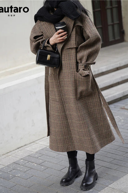 Person wearing a long brown checkered coat holding a coffee cup and black handbag on a city street.