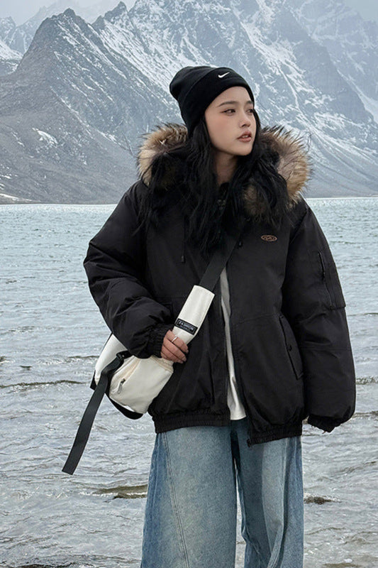 Person wearing a black coat with fur hood standing by a lake with mountains in the background
