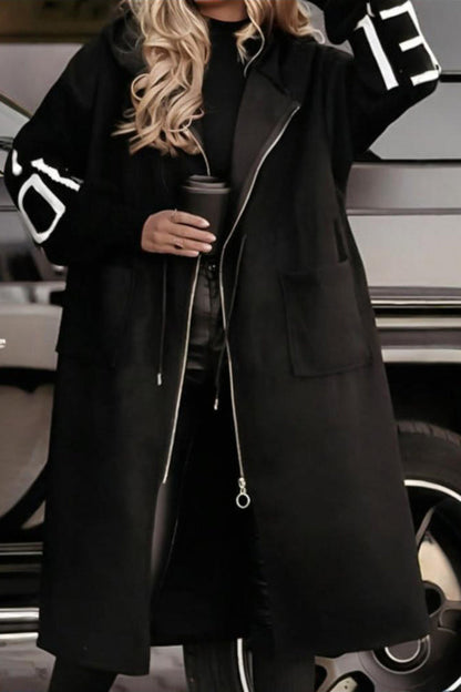 Person wearing a black coat with white lettering, holding a coffee cup, standing next to a car.