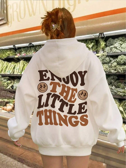 A person facing away from the camera wears a beige oversized hoodie with a slogan 'ENJOY THE LITTLE THINGS' printed on the back, in stylized lettering with cartoon character illustrations.