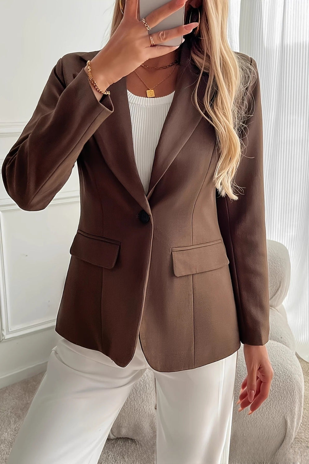 Person wearing a brown blazer over a white top and pants in a room with white walls and curtains.