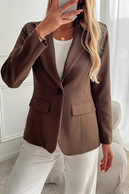 Person wearing a brown blazer over a white top and pants in a room with white walls and curtains.