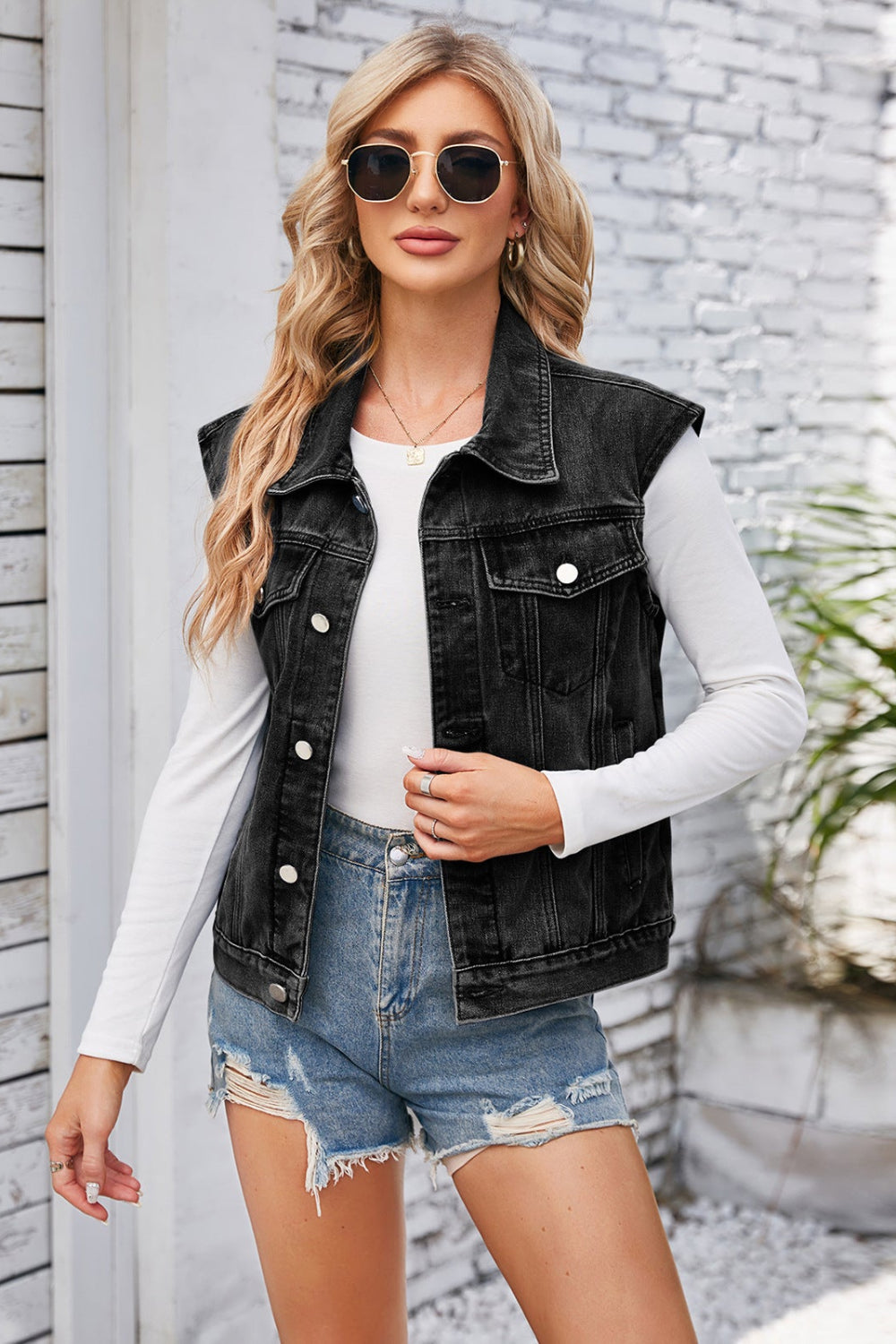 Woman wearing a black denim vest, white long-sleeve shirt, and denim shorts against a light stone wall.