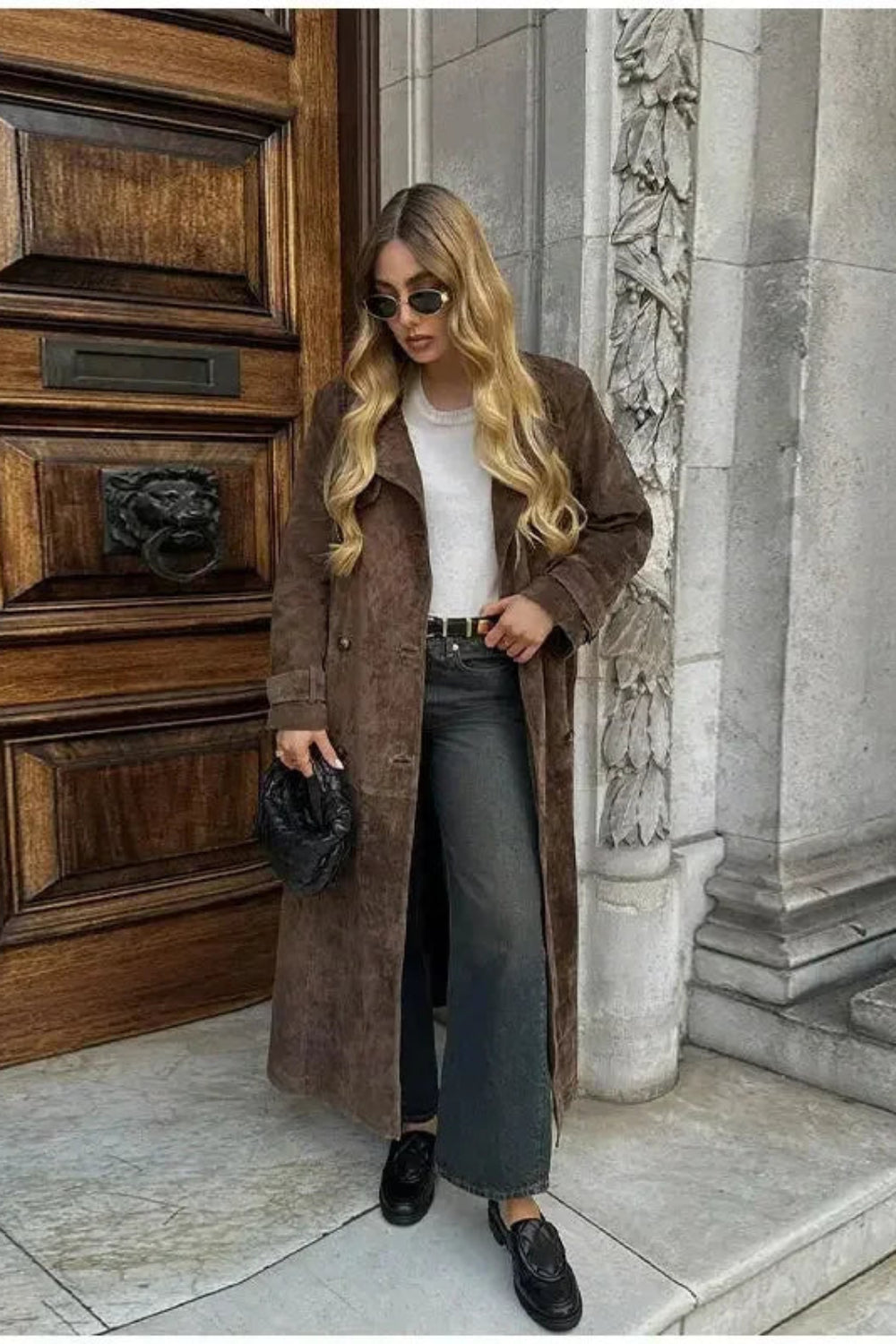 Woman in a brown coat standing in front of a large wooden door.
