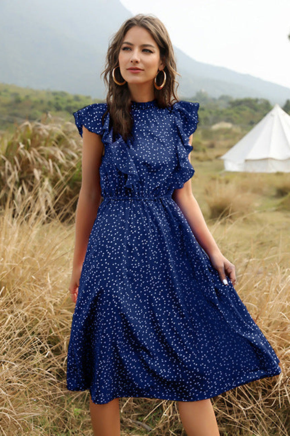 Woman in a blue polka dot dress standing in a field with tents in the background