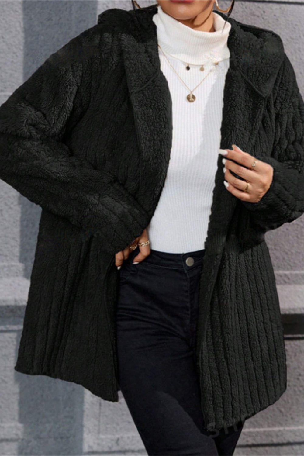 Person wearing a black ribbed knit coat over a white turtleneck and dark pants, standing against a gray stone wall.