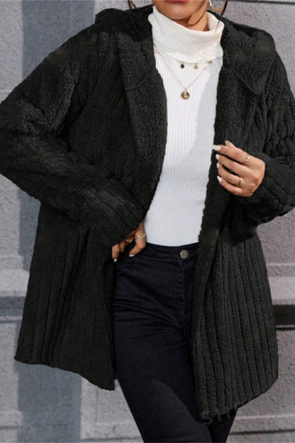 Person wearing a black ribbed knit coat over a white turtleneck and dark pants, standing against a gray stone wall.