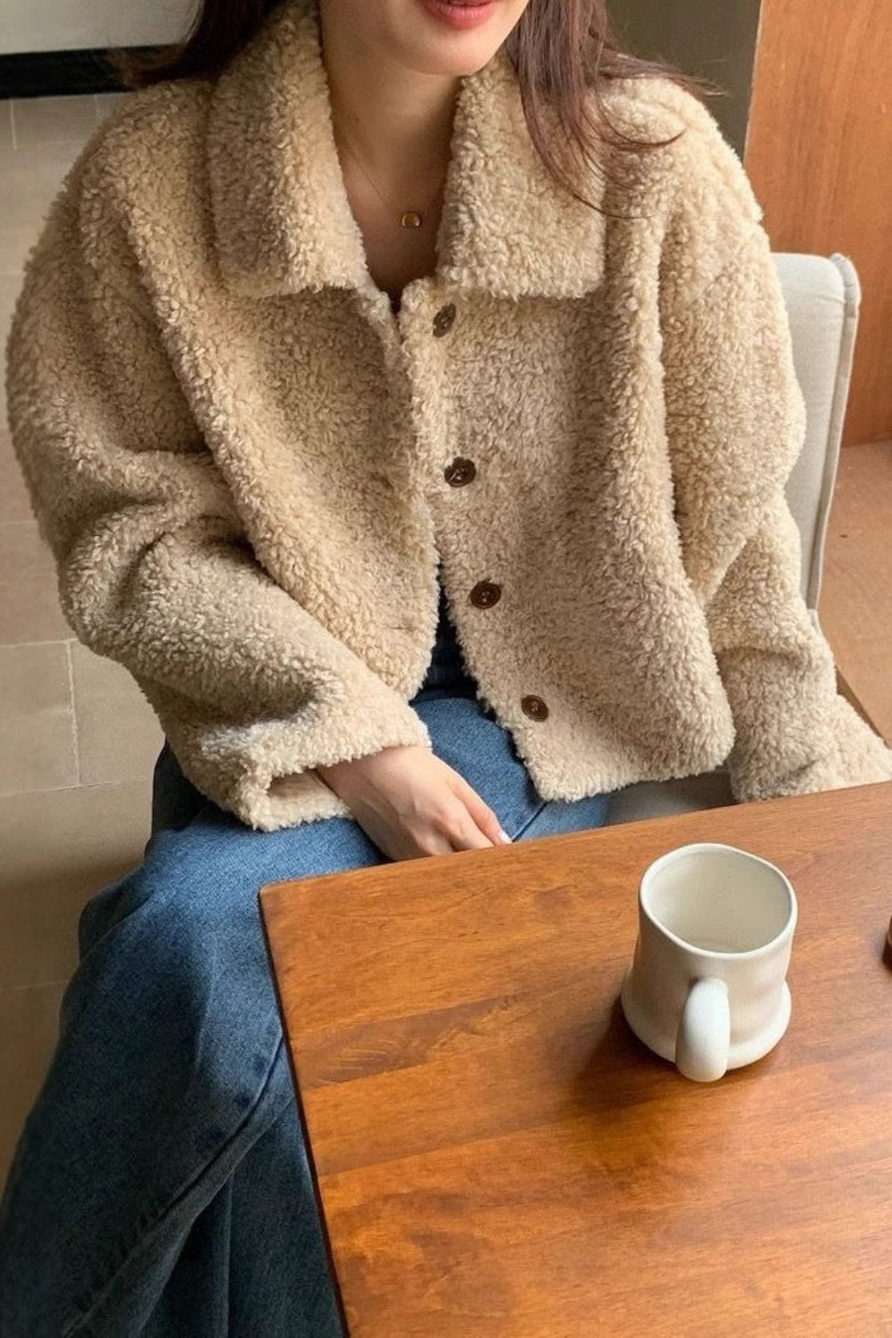 Person wearing a beige sherpa jacket sitting at a wooden table with a mug and small tray.