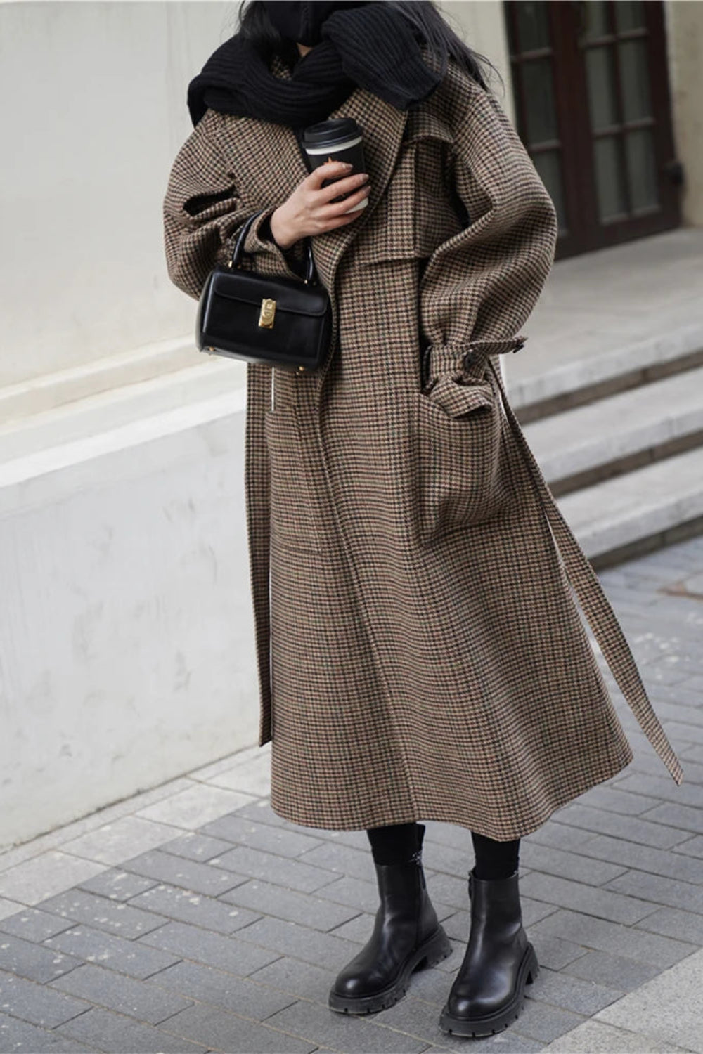 Person wearing a long brown checkered coat holding a black handbag on a city street.