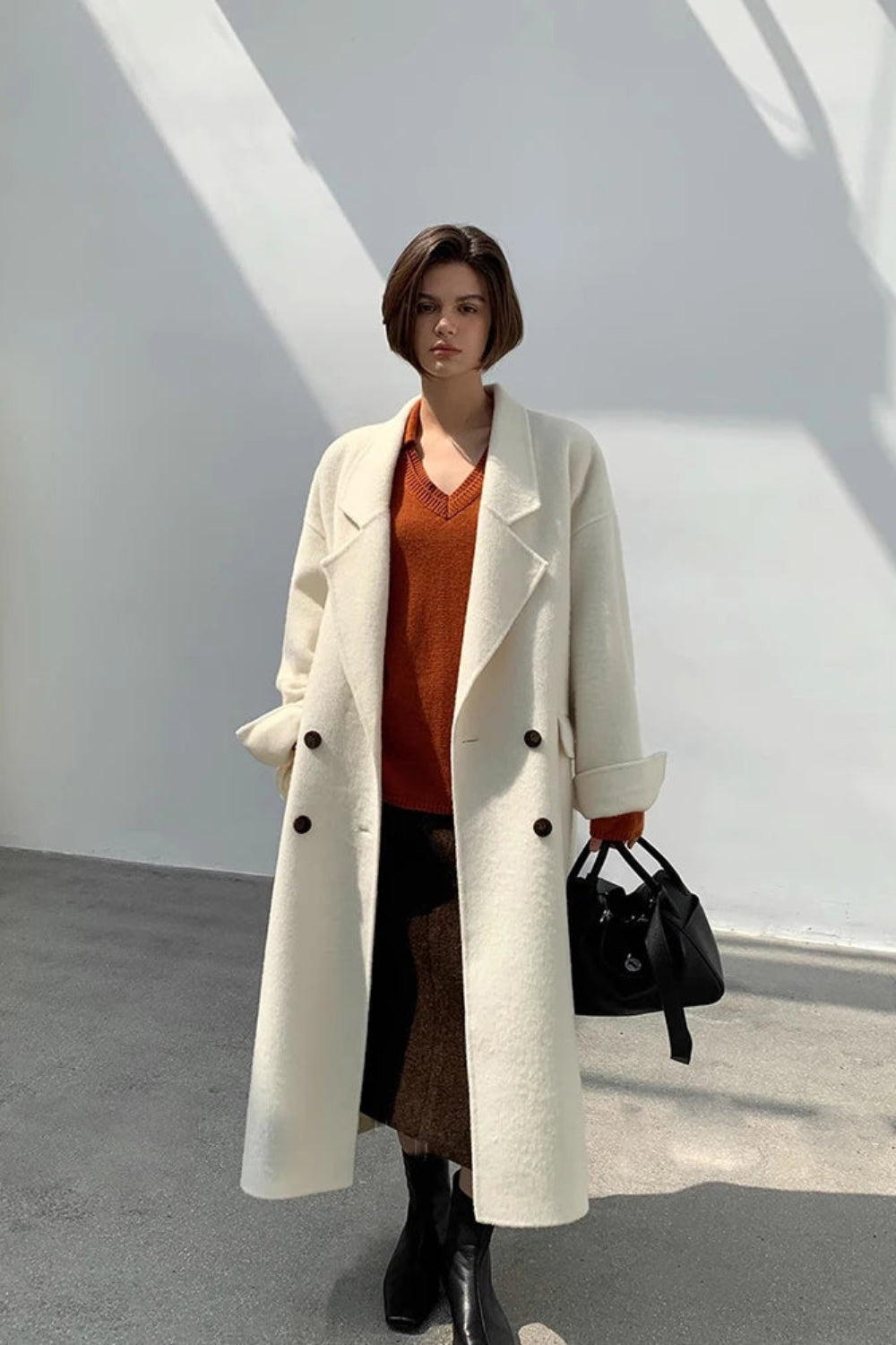 Person wearing a long white coat over a brown outfit, holding a black handbag against a white wall.