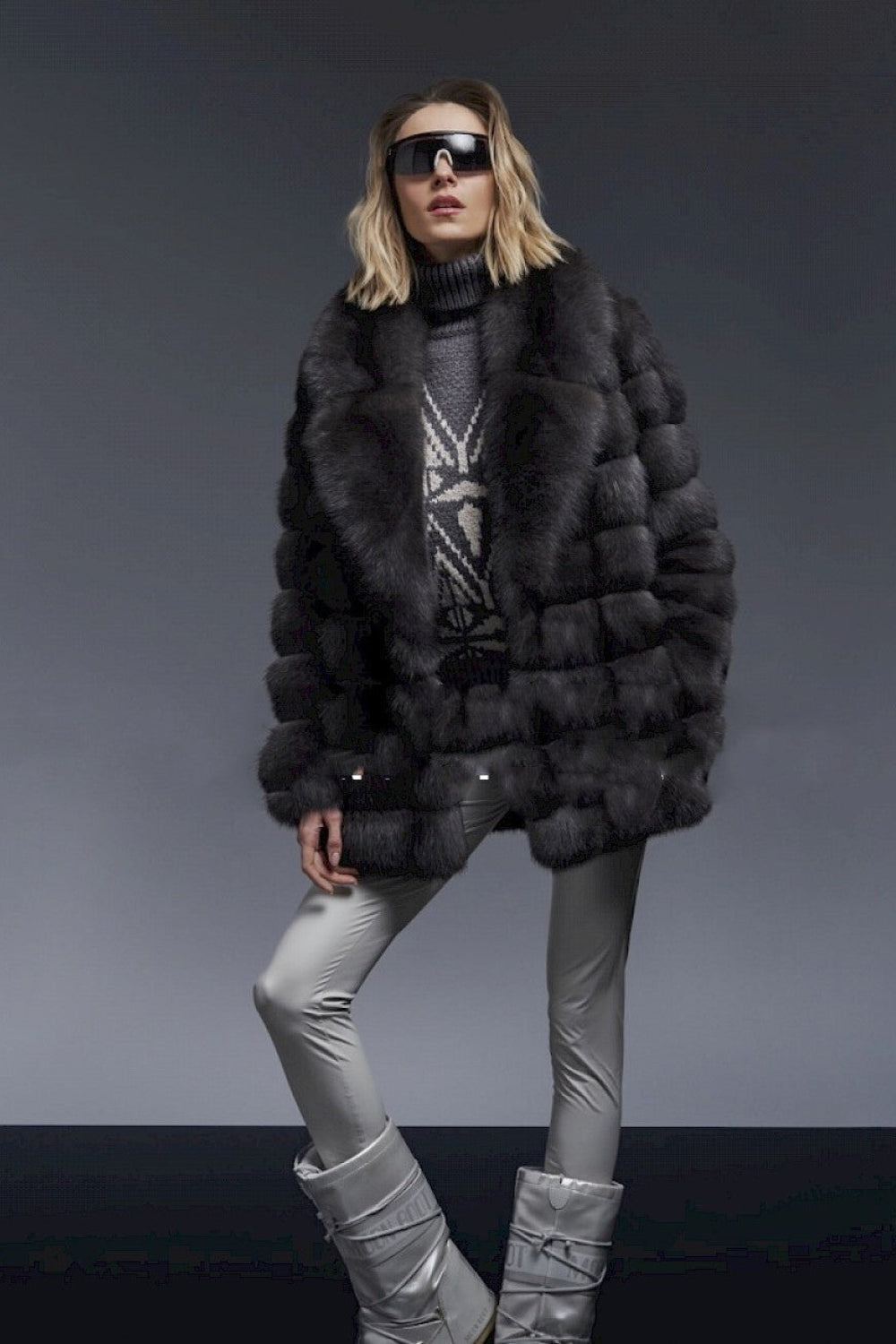 Person wearing a black fur coat, gray sweater, silver pants, and silver boots on a dark background