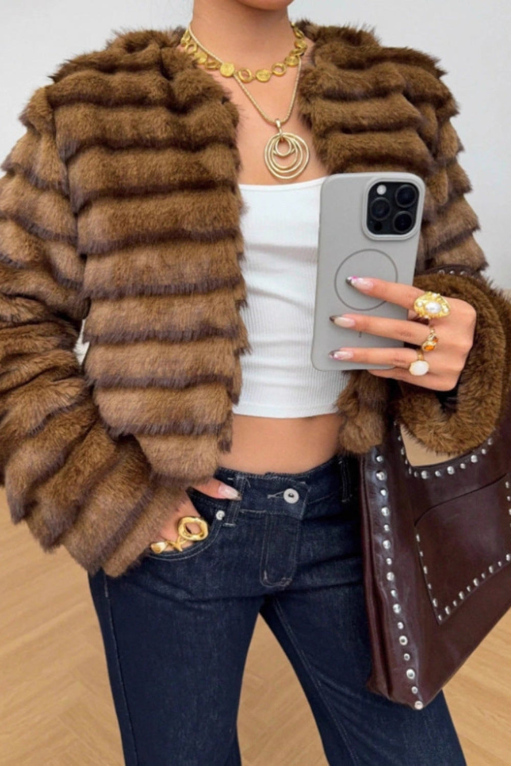 Person wearing a brown faux fur jacket, white top, and dark jeans, holding a phone and a brown handbag.