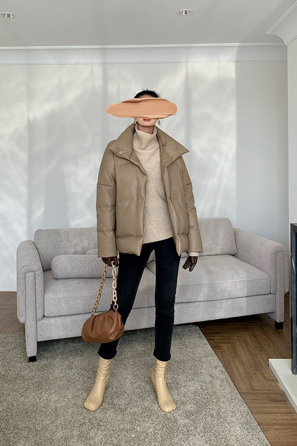 Person wearing a beige jacket, white turtleneck, black pants, and beige boots in a living room.