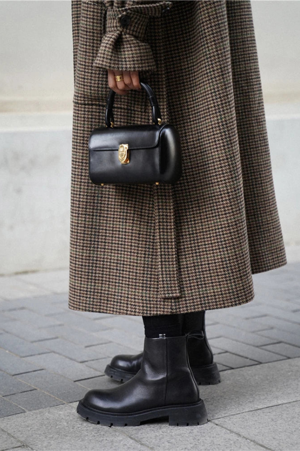 Person wearing a long brown coat and black boots, holding a black handbag on a pavement.