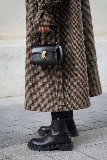 Person wearing a long brown coat and black boots, holding a black handbag on a pavement.