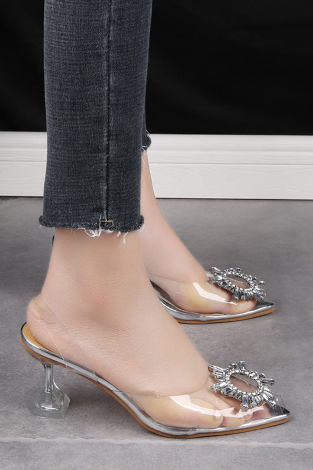 Clear high-heeled shoes with decorative elements worn by a person on a gray floor.