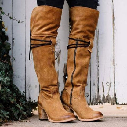 Tan knee-high boots with black straps worn by a person, against a white wooden fence background.