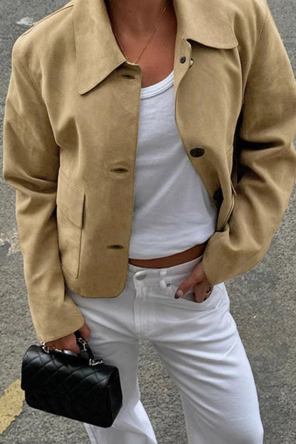 Person wearing a beige jacket, white top, and white pants, holding a black handbag on a street.